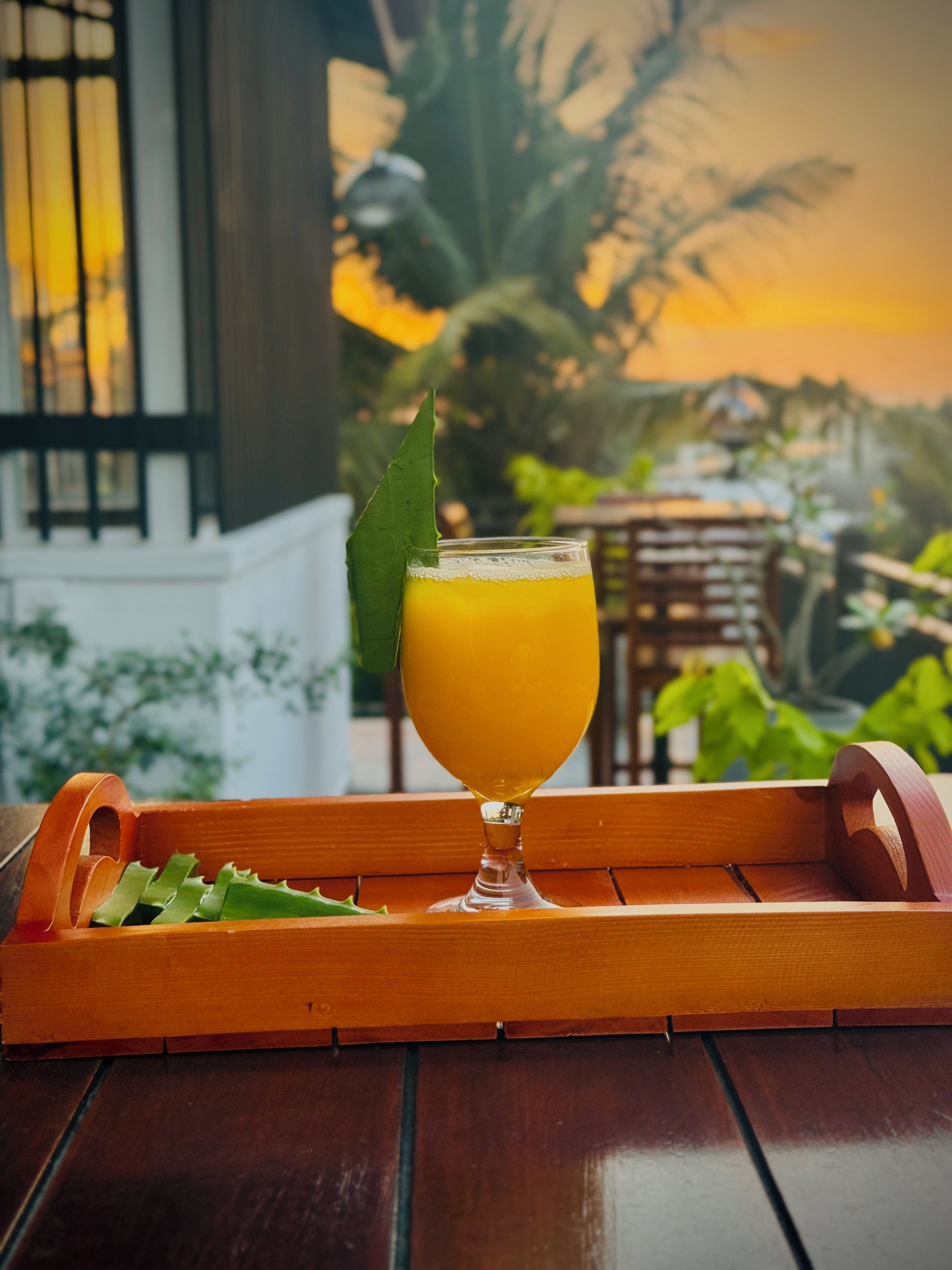A glass of orange juice on a wooden tray with a green leaf garnish, placed on a wooden table with a view of a building and greenery in the background.