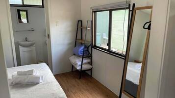 Una habitación pequeña y limpia con una cama, un ventanal, un espejo y un estante con almohadas.