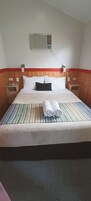Una habitación de hotel con cama, aire acondicionado instalado en la pared, cabecero de madera y detalles en rojo y blanco.