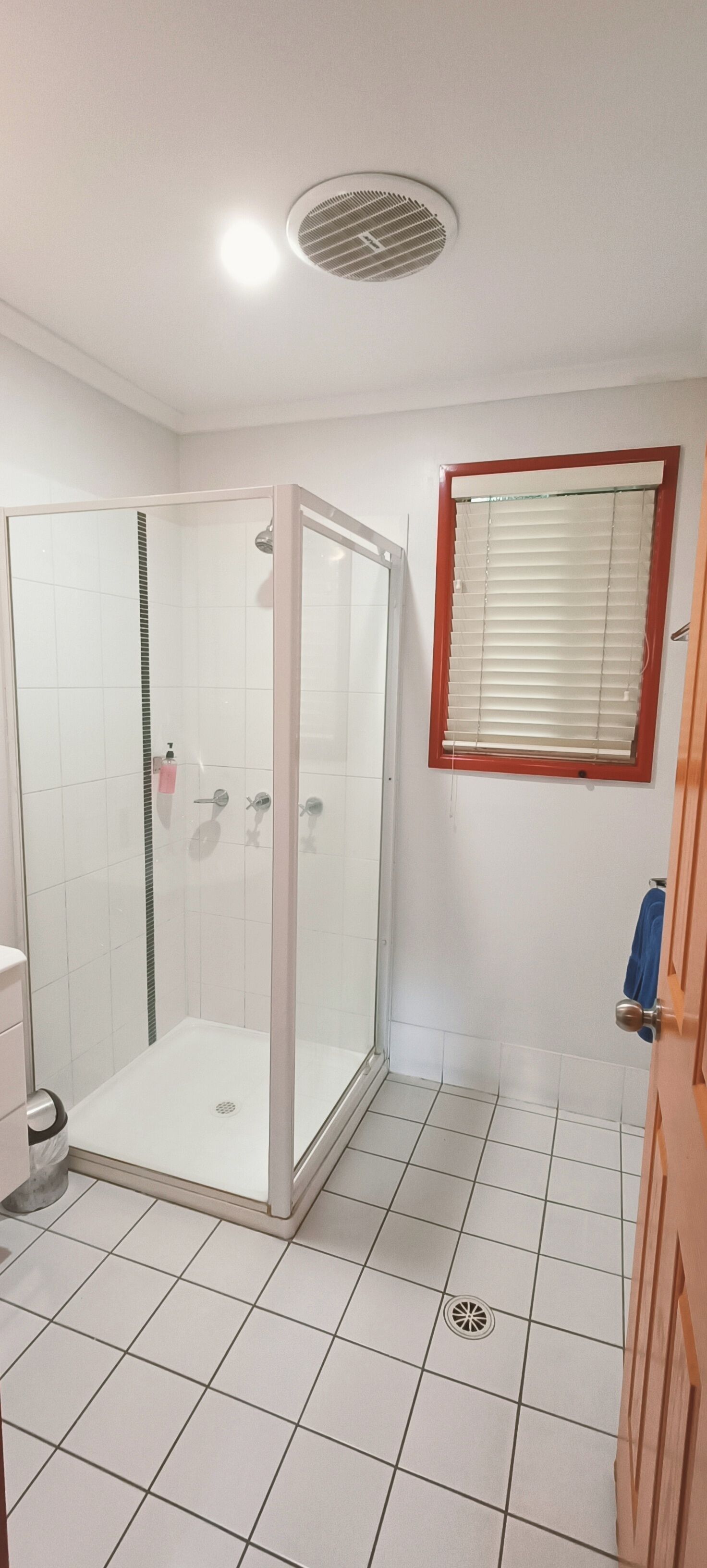 A bathroom with a glass-enclosed shower, white tiled floor, a window with blinds, and a wooden door.