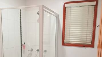 A bathroom with a glass-enclosed shower, white tiled floor, a window with blinds, and a wooden door.