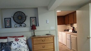 A bedroom with a bed, a wooden dresser, a wall clock, and a kitchen area visible through an open door.