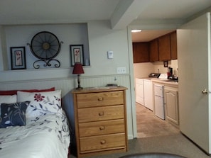 Une chambre à coucher avec un lit, une commode en bois, une horloge murale et un coin cuisine visible à travers une porte ouverte.