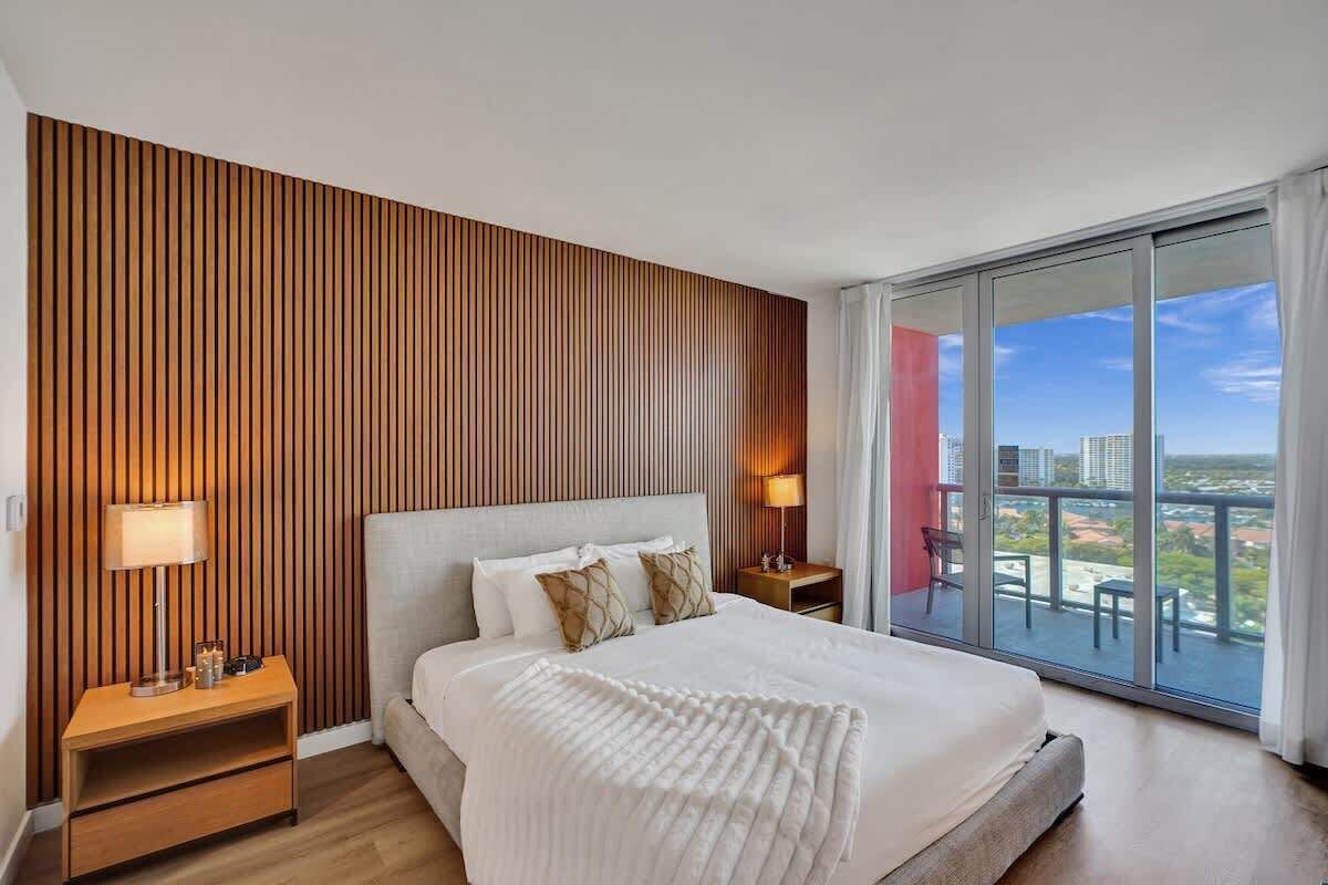 Una habitación de hotel moderna con una cama grande, un panel de pared de listones de madera, un balcón con vistas a la ciudad y una mesita de noche con una lámpara.