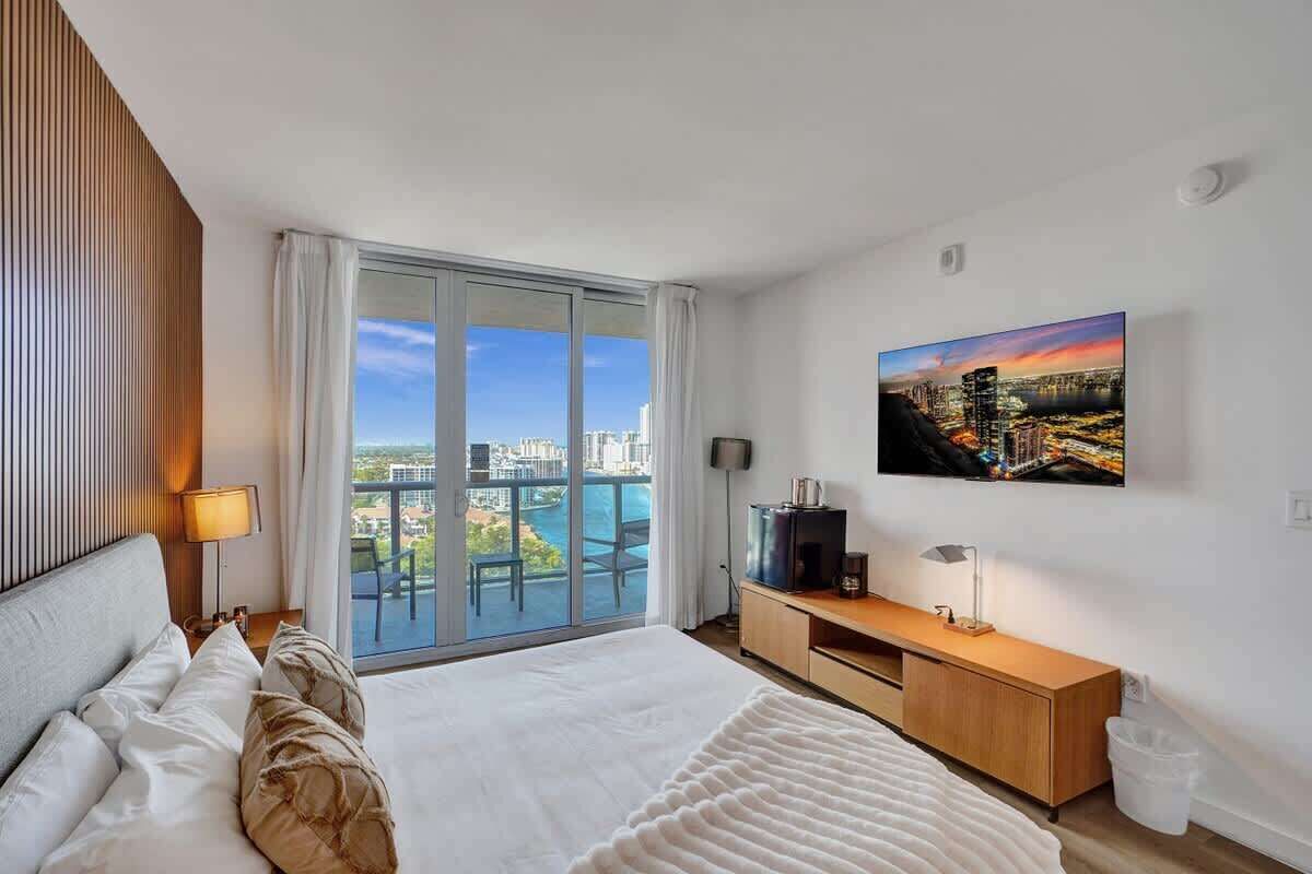 A modern hotel room with a large bed, a TV, a wooden nightstand, and a city view through floor-to-ceiling windows.