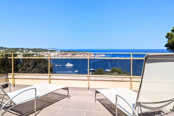 A terrace with two white lounge chairs, a glass balustrade, and a view of the sea with boats.