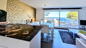 Cocina moderna con una isla grande, zona de comedor con vistas al mar y sala de estar con televisor y asientos.