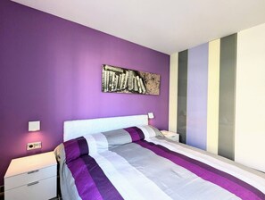 A modern bedroom with a purple wall, a large bed with a striped bedspread, a white headboard, and a wall-mounted artwork.