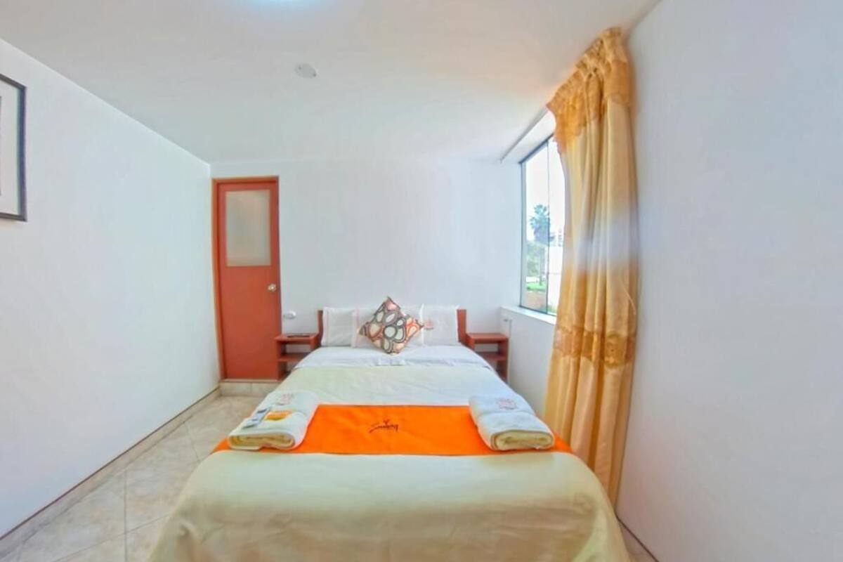 A hotel room with a bed, a window with curtains, a door, and a small table.