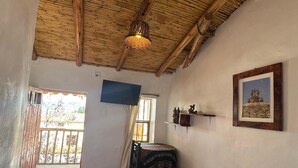 A room with a wooden ceiling, a bed with a quilt, a small table, a chair, a window with curtains, and a picture on the wall. - SUMAQ QUINUA (Carmen Alto)