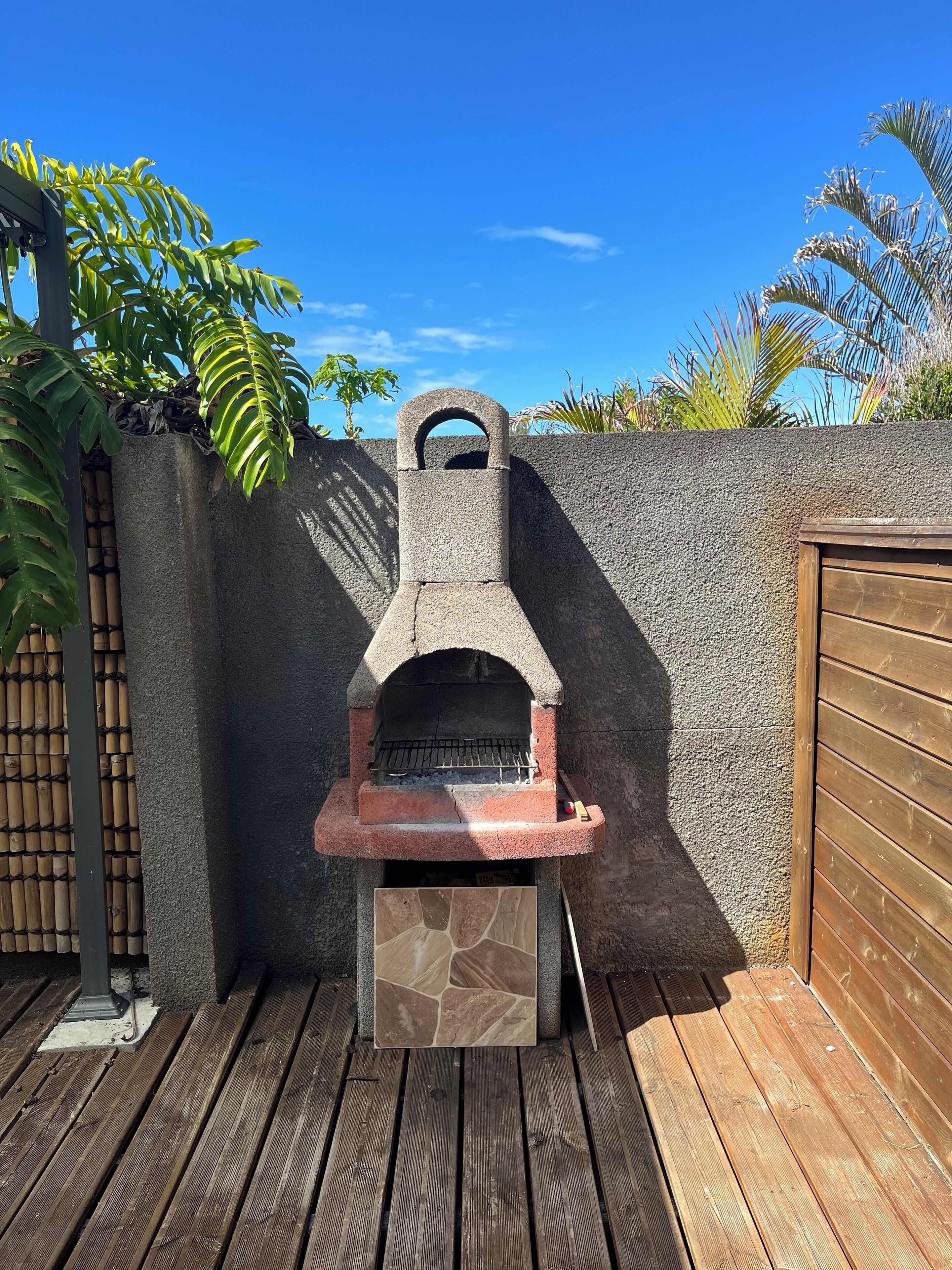 A stone-built outdoor grill with a tiled base, set against a bamboo fence and a wooden deck.
