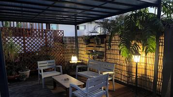An outdoor patio with wooden flooring, two white chairs, a table, and a lamp.