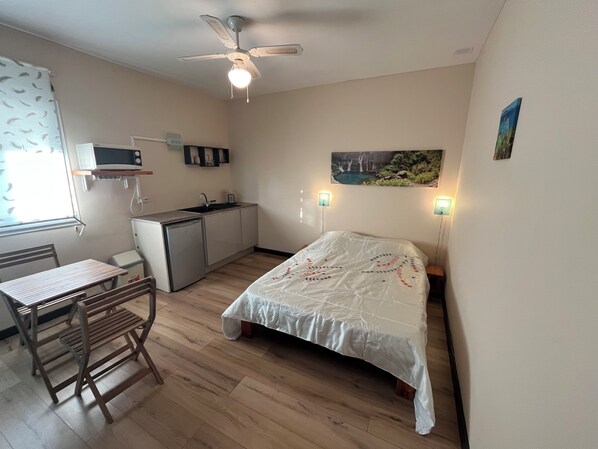 A compact room with a bed, a small kitchenette, a dining table, and a ceiling fan.