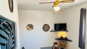 Een moderne woonkamer met een bank, een tafel met glazen blad, een plafondventilator, een aan de muur bevestigde televisie, decoratieve wandkunst en een plank met kleine objecten.