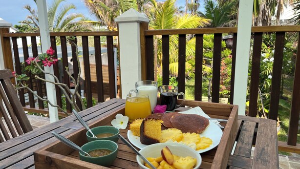 Een houten tafel met een dienblad waarop ontbijtartikelen staan, zoals pannenkoeken, fruit en drankjes, op een balkon met uitzicht op palmbomen en een heldere lucht.