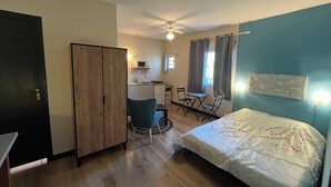 Een kleine kamer met een bed, een bureau, een stoel, een kledingkast en een kitchenette.