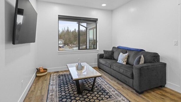A modern living room with a gray sofa, a coffee table, a TV mounted on the wall, and a large window offering a view of trees and houses. - Juneau Family Friendly Getaway 301 (Juneau)