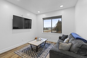 A modern living room with a flat-screen TV, a window with a view, a gray sofa, and a patterned rug. - Juneau Family Friendly Getaway 301 (Juneau)