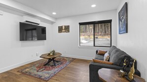 A living room with a sofa, a coffee table, a rug, a painting, and a window with a view of trees. - Accessible, Comfort, Convenient Location 106 (Juneau)