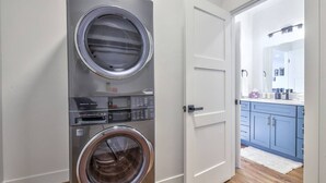 Un cuarto de lavandería con dos lavadoras de carga frontal apiladas y una puerta que da a un baño con espejo y lavamanos.