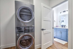 A laundry room with two stacked front-loading washing machines and a door leading to a bathroom with a mirror and sink. - Cozy Retreat With Breath Taking Views 303 (Juneau)