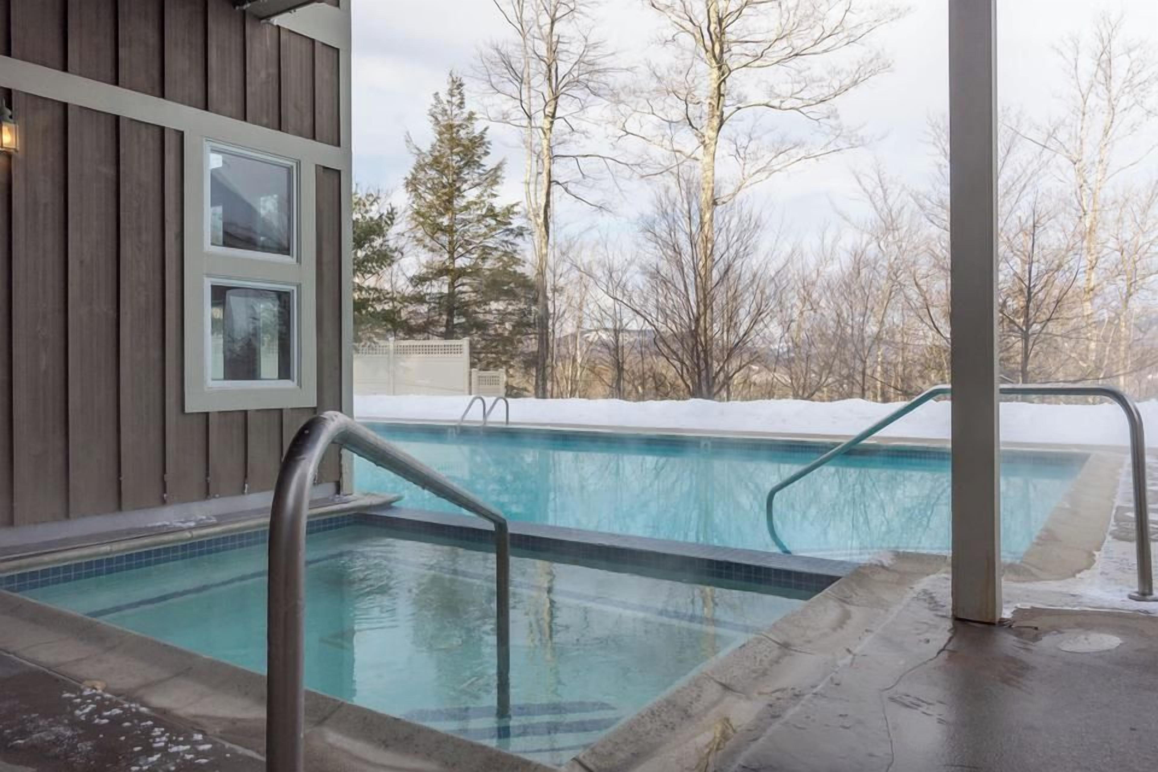 An outdoor swimming pool with a wooden deck and metal railings, surrounded by bare trees and a snow-covered ground.