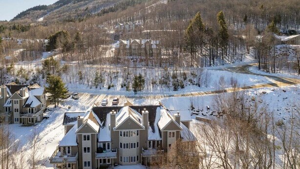 Et snedækket boligområde med huse og en stor bygning, sandsynligvis en lodge eller resort, omgivet af træer og bakker.