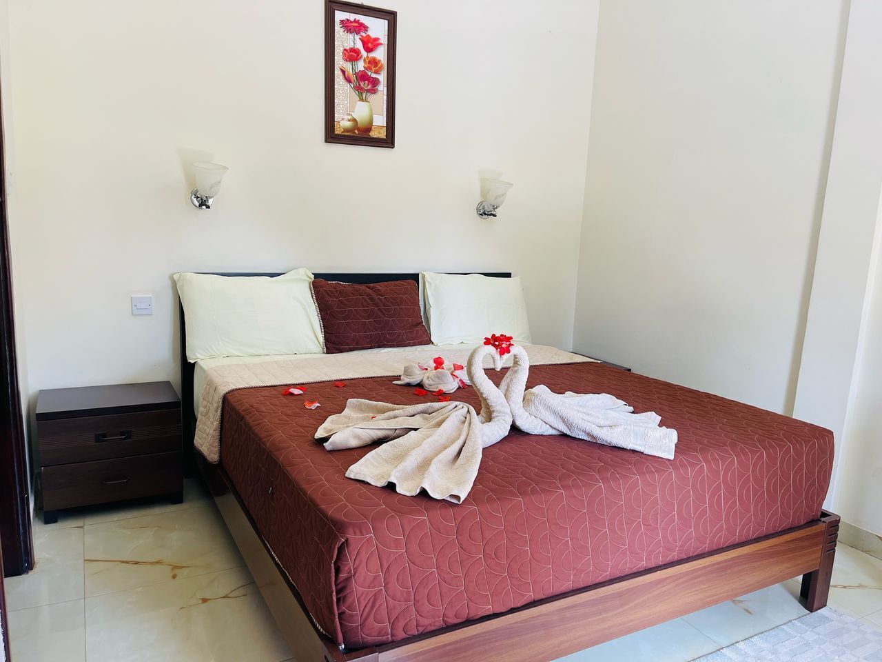 Una cama bien tendida con un colcha roja, una mesita de noche de madera y dos lámparas fijadas a la pared.
