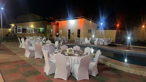 Un espacio de comedor al aire libre con mesas redondas y sillas blancas, una piscina y un edificio con luces al fondo.
