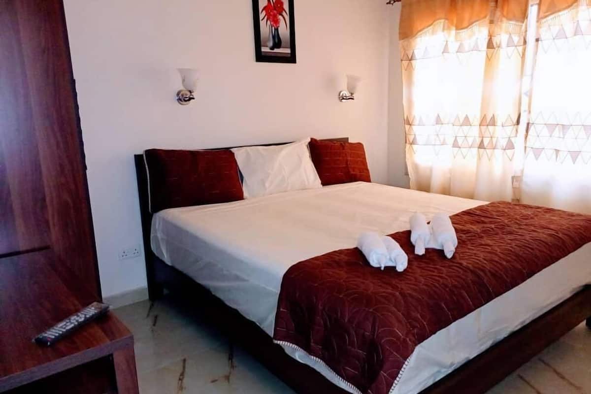 A hotel room with a large bed, two wall-mounted lights, a painting, and a window with patterned curtains.