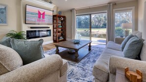 Amplia sala de estar con un televisor grande, asientos cómodos, una mesa de centro de madera y vistas a un jardín a través de amplios ventanales.