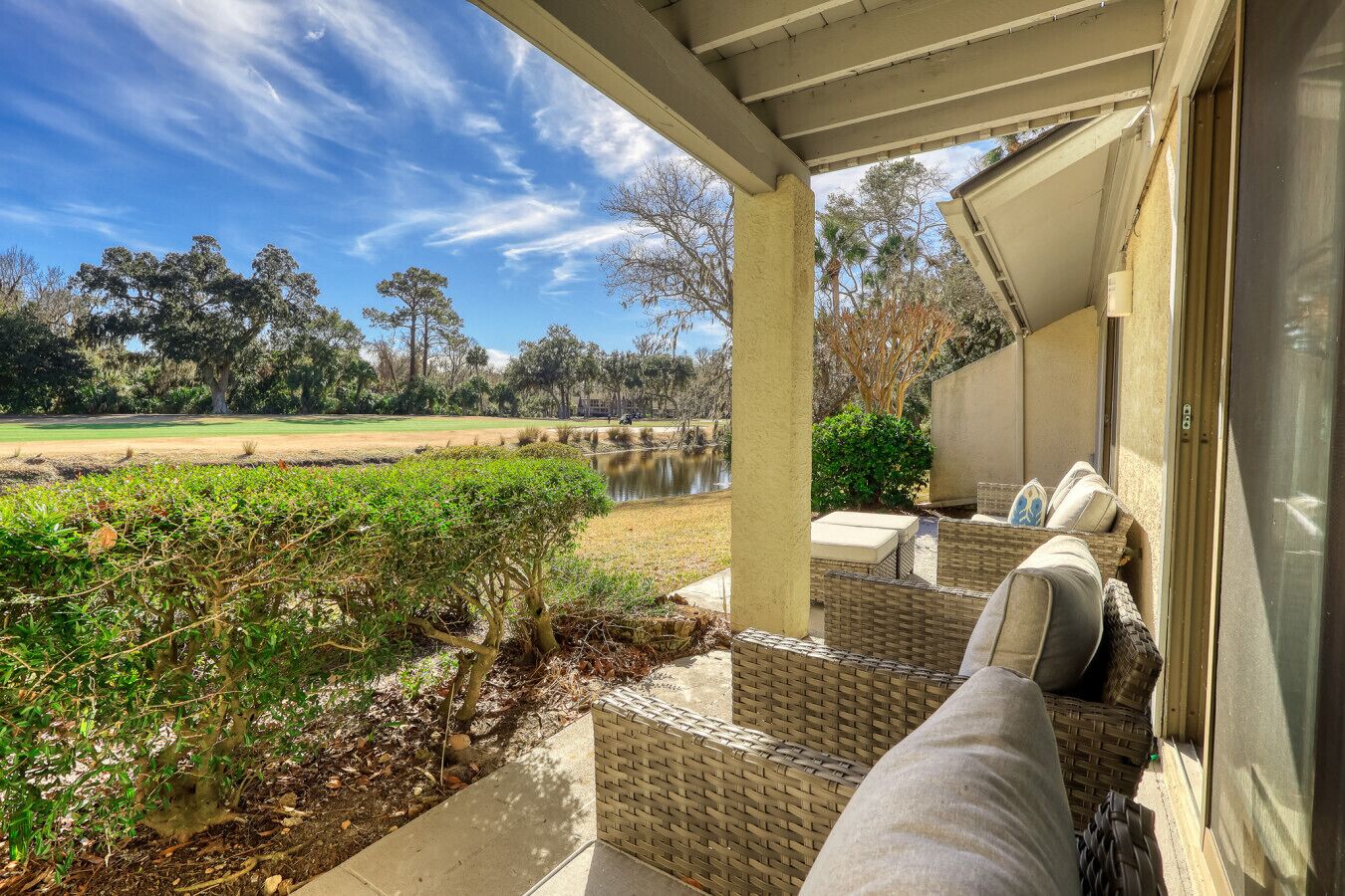 A covered outdoor seating area with wicker furniture and cushions, overlooking a pond and a golf course.