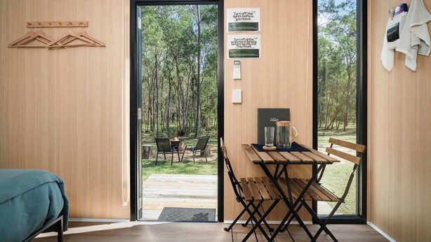 A modern room with a wooden interior, a table with chairs, a bed, and a view of a forest through large glass doors.