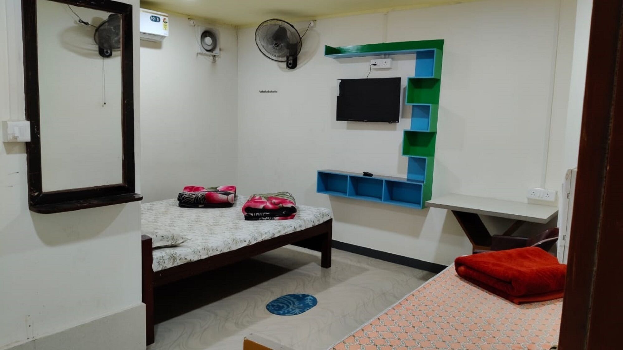 Une petite pièce bien éclairée, équipée d’un lit, d’un miroir, d’un téléviseur fixé au mur, d’un ventilateur et d’une étagère avec des boîtes de rangement.