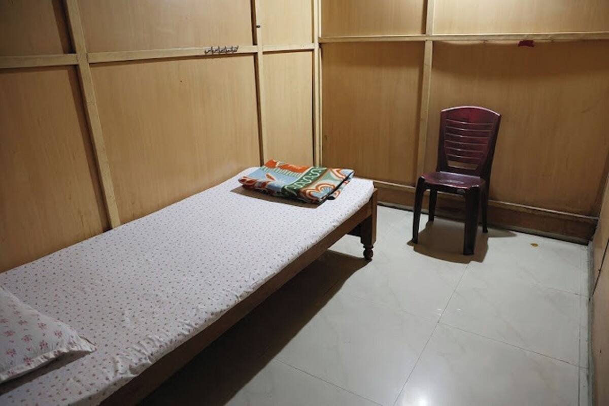 A small, single-bed room with a wooden bunk bed structure in the background, a chair, and a folded blanket on the bed.