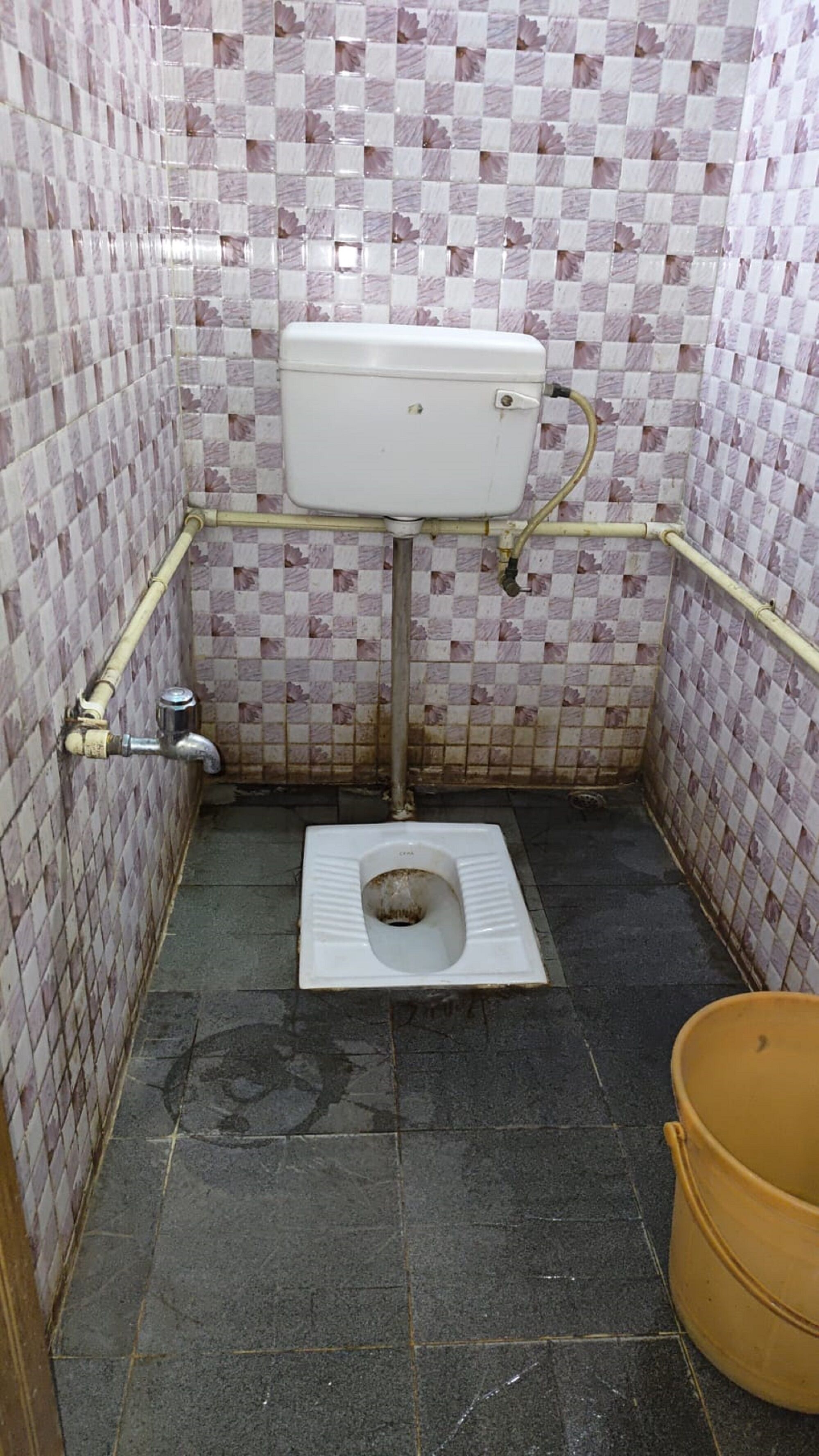 Een kleine betegelde badkamer met een toilet, een gele emmer en een waterreservoir aan de muur.