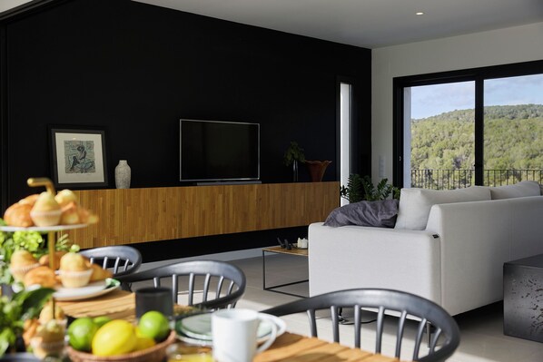 Una sala de estar moderna con un ventanal amplio, un televisor de pantalla plana y una zona de comedor con una mesa y sillas de madera.