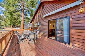 A wooden deck with a table and chairs, a large sliding glass door, and a view of a forested area.