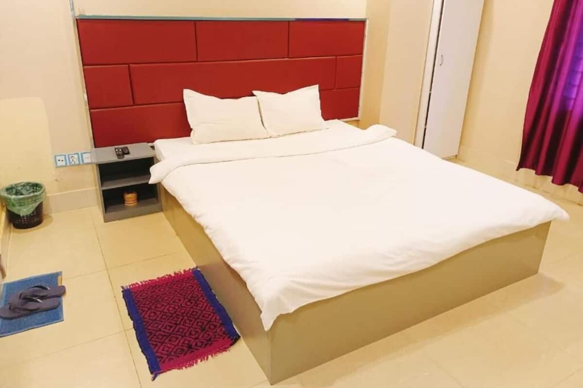 Une chambre d’hôtel avec un grand lit, une tête de lit rouge, une petite table de chevet et un tapis rouge et bleu sur le sol.