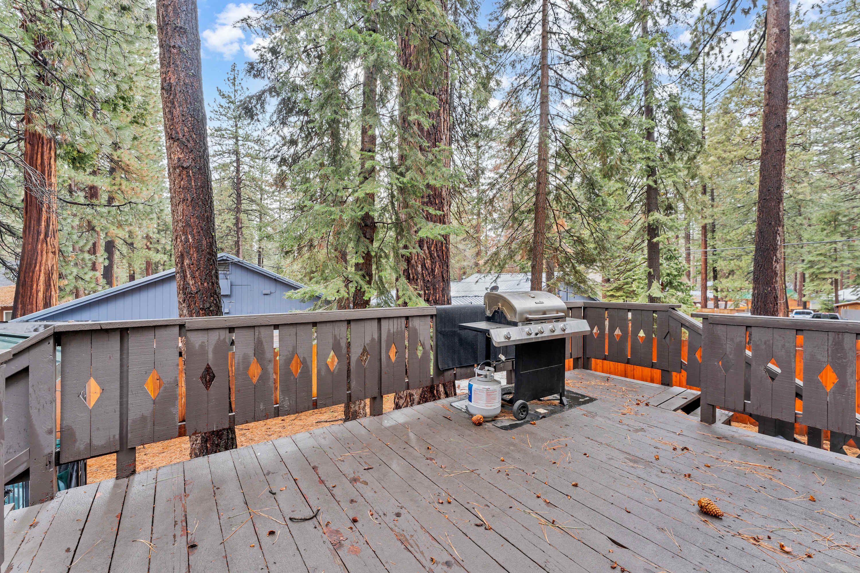 Een houten terras met een barbecue en propaantank, omgeven door hoge bomen.