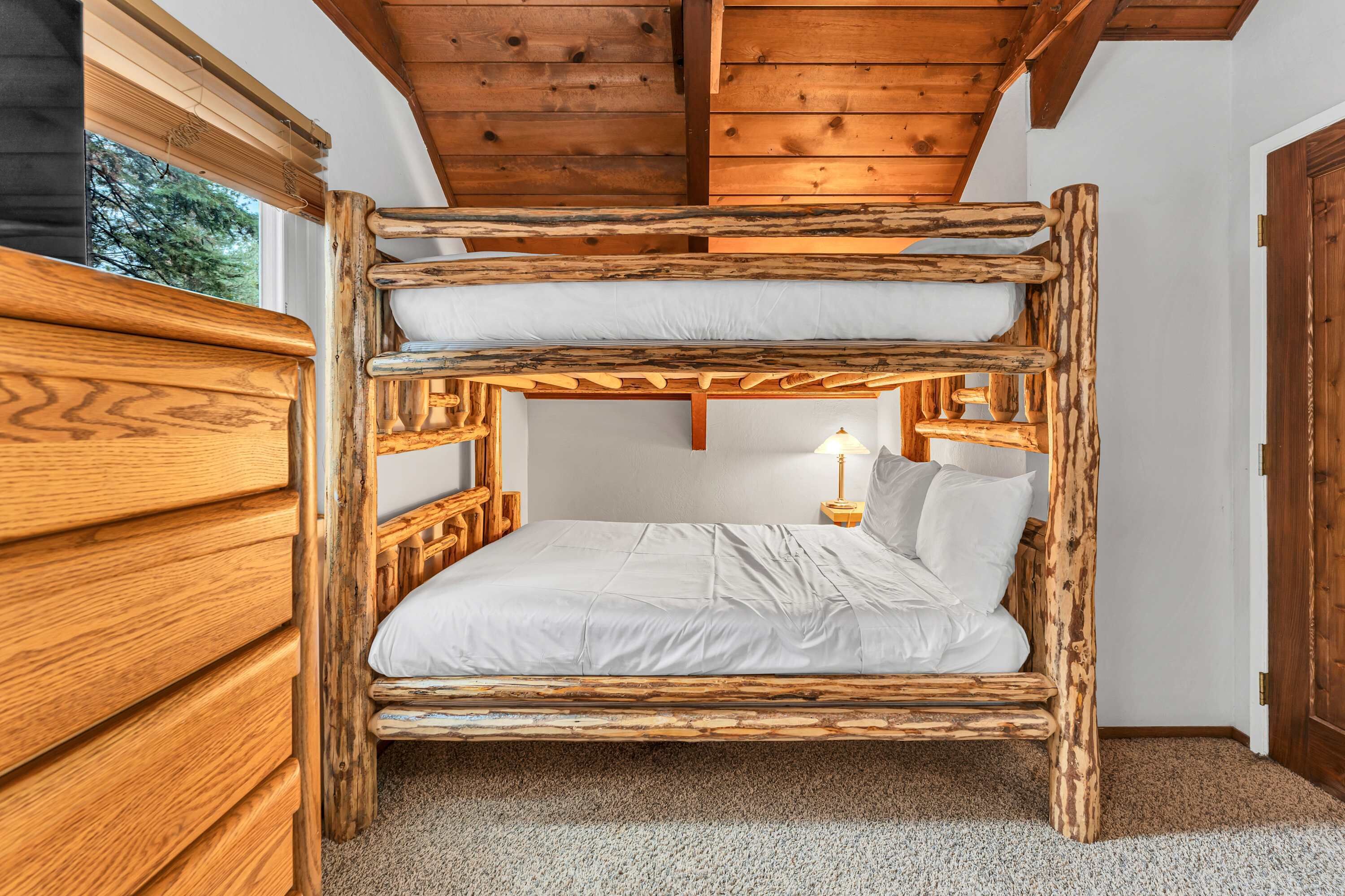 A bunk bed with a wooden frame and white bedding, a wooden dresser to the left, and a window with blinds.