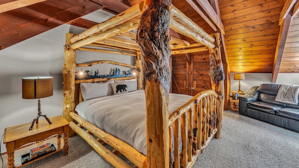 A rustic wooden four-poster bed with a bear-themed headboard, a bedside table with a lamp, and a leather sofa in a cabin-style room.