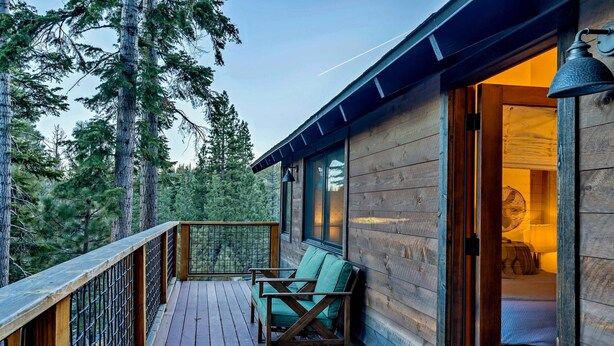 Eine Holzterrasse mit einem Stuhl, ein Bett ist durch eine offene Tür sichtbar, und es bietet Blick auf hohe Bäume.