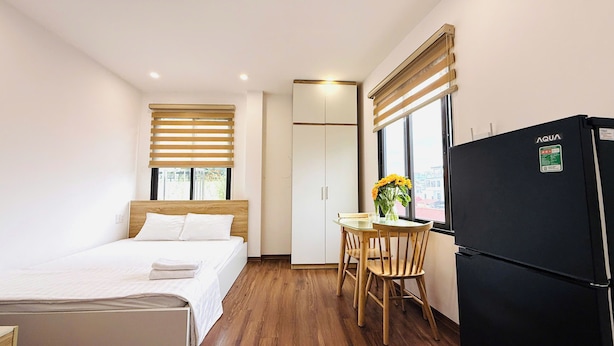 Une chambre d’hôtel avec un lit, une petite table sur laquelle est posé un vase de fleurs, un réfrigérateur et une fenêtre avec des stores.