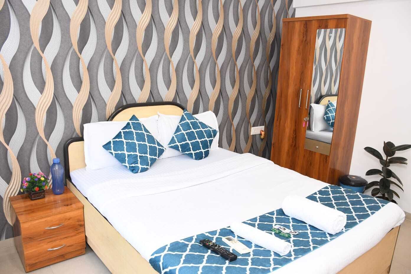 Una cama bien hecha con ropa de cama de diseño azul y blanco, una mesita de noche de madera con un jarrón y flores, y un mueble espejo.