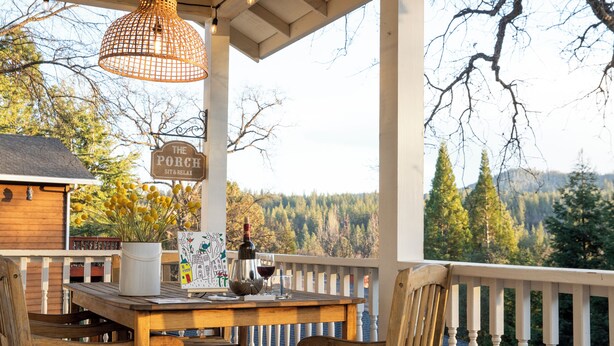 Et træbord med stole på en veranda, et skilt med teksten "The Porch Sit & Relax", en potteplante, en vinflaske og glas.