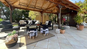 Un espace extérieur aménagé, abrité et doté d’un plafond en bois, d’un canapé, de deux fauteuils, d’une table et de plantes en pot.