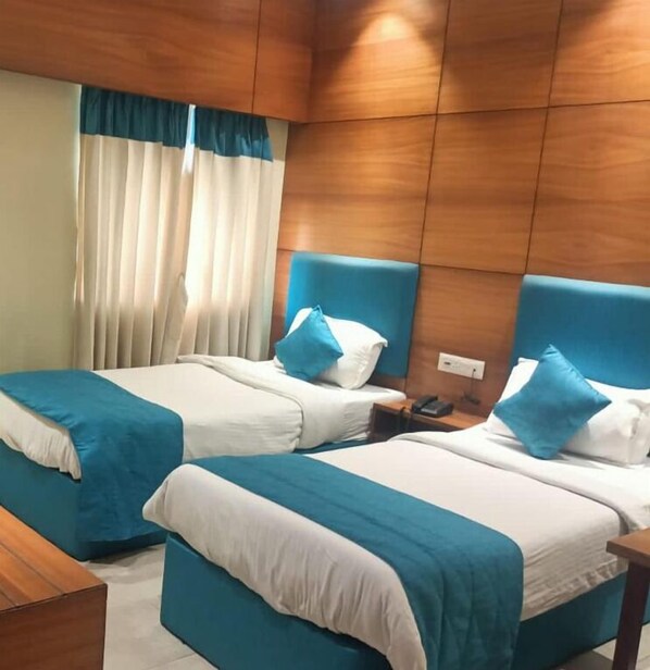 A hotel room with two beds, each with a blue headboard, white bedding, and blue and white striped blankets. There is a wooden headboard, a nightstand with a phone, and a window with curtains.