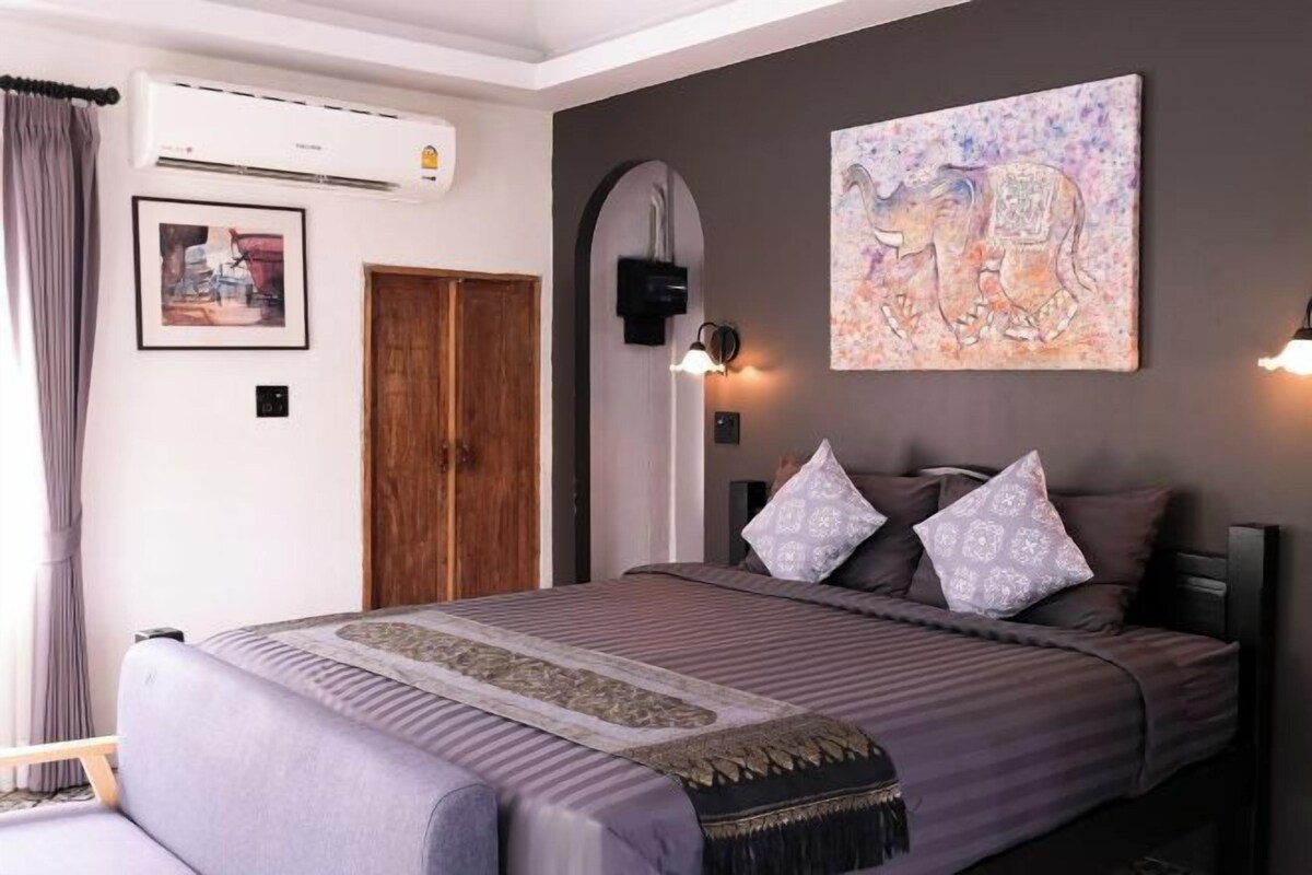 A bedroom with a bed, a wall-mounted air conditioner, a framed picture, a door, and a wall painting.