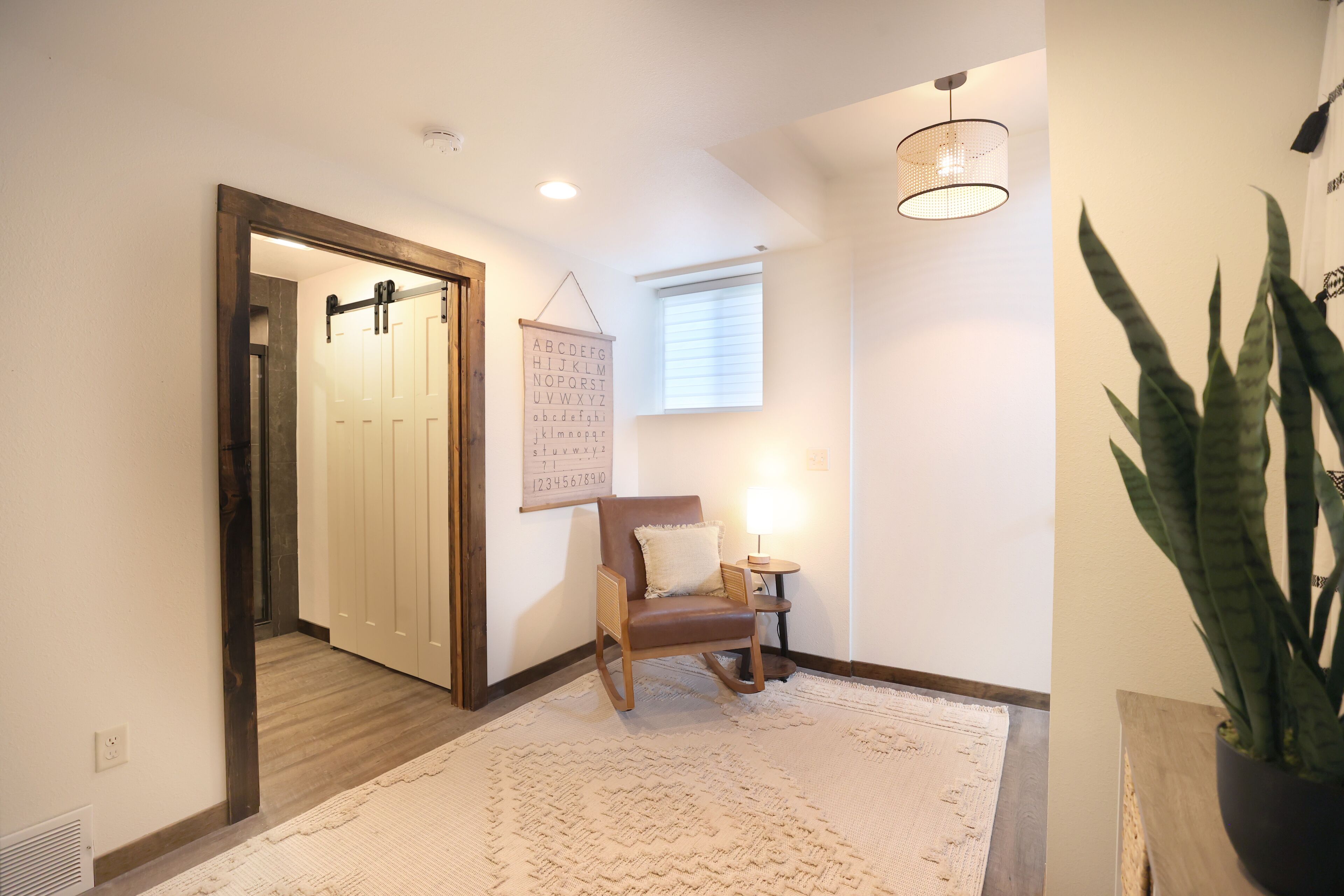 A hallway with a wooden door, a rocking chair, a small table with a lamp, a potted plant, and a wall-mounted alphabet chart.